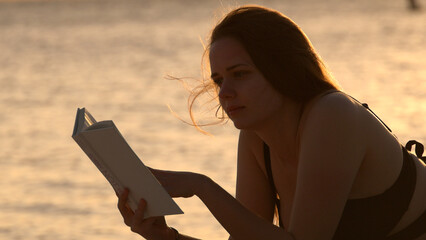 Young woman reads a book while lying on the beach at sunset