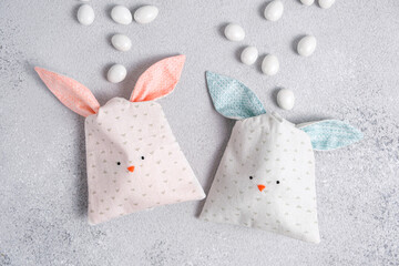 Two bag of sweets in shape of Easter bunny with blue and pink ears on light gray background. Packing of cotton fabric in form of cute bunnys or rabbits, sewing for children. Scattered chocolate eggs.