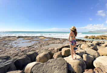 Woman standing on rocks and watching the sea in Hawaii. Young model with head cap on her back