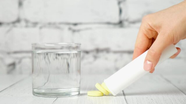 clear glass of water and tube of yellow soluble round effervescent tablets on white background. A woman's hand takes out liquid vitamins in close-up. Treatment and prevention of viral diseases.