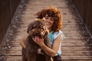 Curly-haired woman playing with her brown spanish water dog sitting on the ground on a wooden walkway. Lifestyle