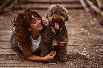 Curly-haired woman playing with her brown spanish water dog sitting on the ground on a wooden walkway. Lifestyle