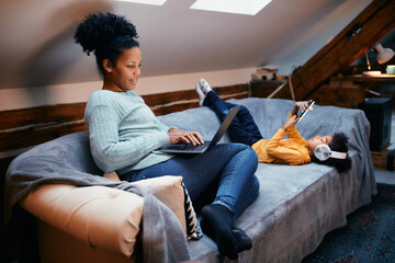 Happy black woman works on laptop while daughter using using smart phone at home.