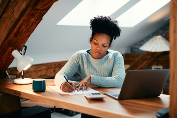 African American woman taking notes while working on laptop at home.
