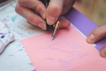 Naklejka premium Process of calligraphy handwriting with an ink fountain pen feather, calligrapher practicing writing on a postcard paper using pen brush and sign pen with inkwell on master-class lesson, hands view