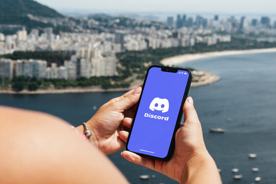 Girl holding smartphone with Discord app on screen. City and bay with some boats in the background. Rio de Janeiro, RJ, Brazil. March 2022