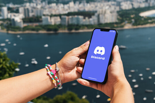 Girl holding smartphone with Discord app on screen. City and bay with some boats in the background. Rio de Janeiro, RJ, Brazil. March 2022