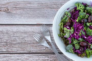 Fresh colorful vegetable salad bowl with red cabbage, broccoli, olives, and lettuce with forks on wooden table with copy space. The concept of healthy plant-based eating.