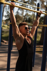 Obraz premium trendy handsome sports man with sunglasses in a stylish black T-shirt trains, workout and pulls up in the park. Healthy lifestyle