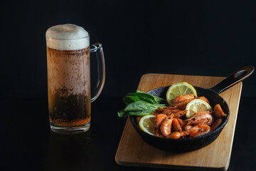 on a dark background there is a glass with beer filled with a foamy drink and on a wooden board there is a frying pan with ready-made shrimps and lemon