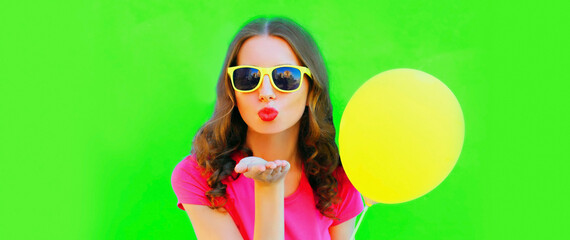 Portrait of beautiful young woman blowing her lips sending air kiss with yellow balloon on green background