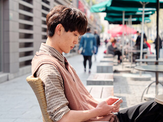 Portrait of handsome Chinese young man with curly black hair in plaid shirt and wool vest sitting outdoor cafe with his mobile phone in hand in sunny day, male fashion, cool Asian young man lifestyle