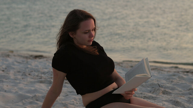 Young woman reads a book while lying on the beach at sunset