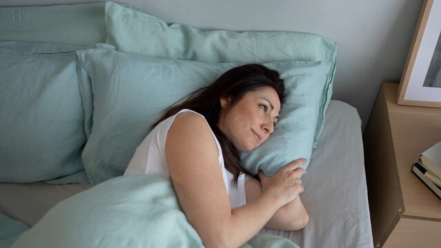 Top view portrait of a smiling woman in a comfortable bed with blue linen sheets, pensive and happy resting, waking up in the morning after a good sleep in a cozy home or hotel room