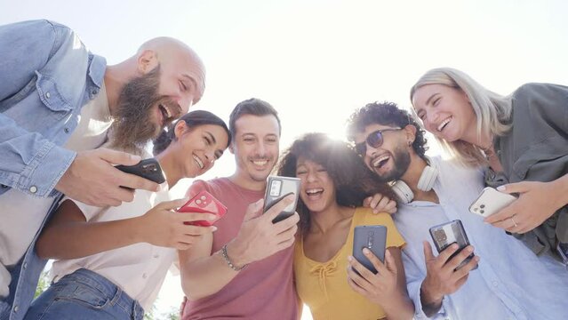 Low angle of Multiracial happy group of friends using phone and smiling together outside. People addicted to technology and social media