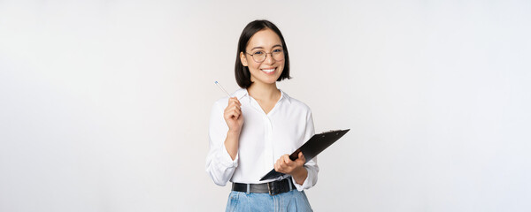 Smiling young asian woman taking notes with pen on clipboard, looking happy, standing against white...