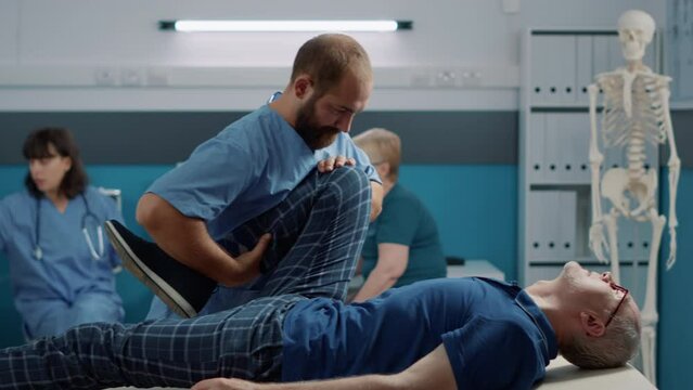 Male Osteopath Cracking Legs Bones And Helping Old Man With Muscles Stretch At Physical Therapy Appointment. Osteopathy Specialist Practicing Alternative Medicine On Senior Man.