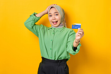 Amazed young Asian woman in a green shirt showing credit card on yellow background
