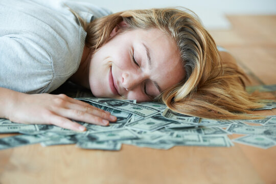 For the love of money. A smiling girl with her eyes closed lying down on the floor with her head in a pile of money.