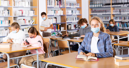 Group of intelligent girls and boys in face masks preparing for exams in library. Teenager girl looking in camera.