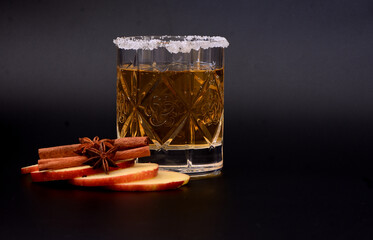 Cider in a crystal glass with sugar on the rim, cut red apples, anise and cinnamon sticks on a black background.