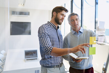 They believe in constant innovation to impress their clients. Shot of two male coworkers arranging sticky notes on a glass wall during a brainstorming session.