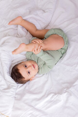 cute smiling baby in a green cotton bodysuit is lying on a white bed on his back. top view