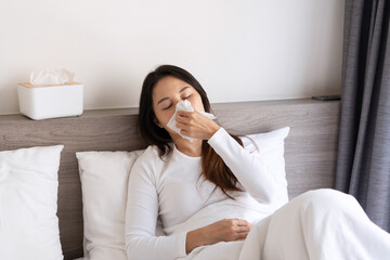 Sick young Asian woman feeling cold and has a high fever blowing nose while lying down on bed in the morning at home. Allergy, flu concept. - obrazy, fototapety, plakaty
