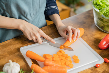 Asian young woman, girl or housewife using knife, cutting carrots on  board, on wooden table in kitchen home, preparing ingredient, recipe fresh vegetables for cooking meal. Healthy food people.