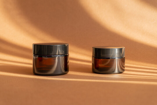Two jars of amber glass for cosmetics on brown background with shadow in form of stripes. Mock-ups of containers with moisturizer for face, hands, close-up. Concept of body care