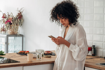 Fototapeta na wymiar Young mixed race woman using mobile phone to send text message at home kitchen. Copy space.