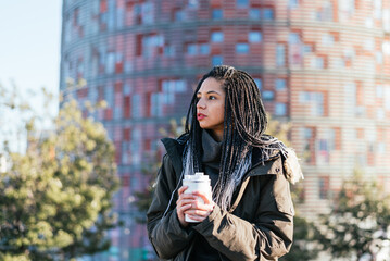Pensive Hispanic female in warm outerwear with dreadlocks and to go thermos mug with hot beverage...