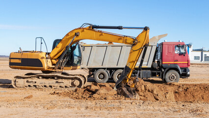 A powerful crawler excavator loads the earth into a dump truck against the blue sky. Development and removal of soil from the construction site.