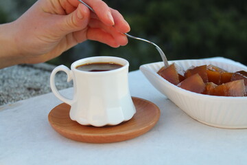 Cup of coffee, a woman take a piece of pumpkin dessert (known as “kirecte kabak tatlisi” in Hatay, Turkey) with fork from white plate in balcony. Selective focus