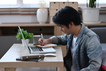 Concentrated male freelancer working online with laptop computer and making notes on notebook.