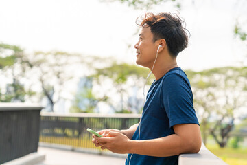 Asian man listening to music from earphones and smartphone application while jogging at city park i summer morning. Healthy male athlete enjoy outdoor sport training workout exercise and running