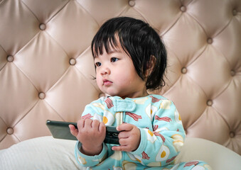 Little child sitting on the bed and playing with smartphone . Little baby girl using smartphone at home. Excited Asian little girl is using a smartphone. People using technology.