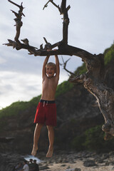Boy child on the ocean is pulled up on a tree branch.