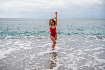 A beautiful and sexy brunette in a red swimsuit on a pebble beach, Running along the shore in the foam of the waves