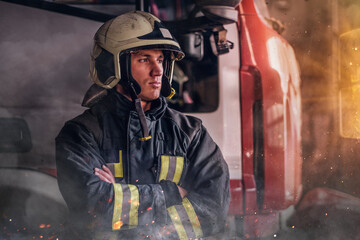 Fireman wearing protective uniform standing next to a fire engine in a garage of a fire department, crossed arms and looking sideways