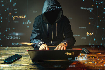 A hacker anonymous in a hood is typing on a laptop keyboard in a dark room. Cybercrime fraud and...
