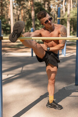 Handsome athlete man with naked muscular body and tattoo in sunglasses and headphones doing stretching in the park
