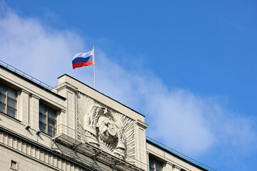 Russian flag on the parliament building in Moscow on background of blue sky and white clouds. Facade of State Duma of Russia with soviet coat of arms, russian authority