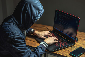 Hacker in a hood with a phone is typing on a laptop keyboard in a dark room. Cybercrime fraud and...