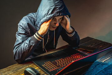 Hacker in a hood with a phone is typing on a laptop keyboard in a dark room. Concept of cyber warfare and Dos attacks - obrazy, fototapety, plakaty