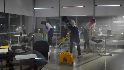 Multiethnic janitors in safety mask mop floor and disinfect furniture in empty office