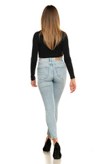 Rear view of a young woman walking in studio in black short blouse and blue jeans on a white...