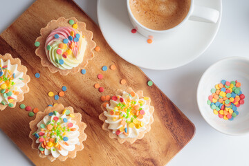 Top view of mini cupcakes decorated with candy sprinkles and cup of coffee on wooden background.