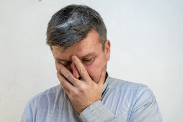 A mature man covers his face with his hand against the light wall. A male with a short haircut and graying hair The emotion of grief, of loss