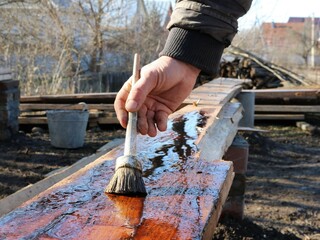 processing wood with dark technical oil outdoors, a man's hand with a round brush applies oil impregnation on a wooden board in a backyard in a rural area, wood impregnation with dark oil liquid